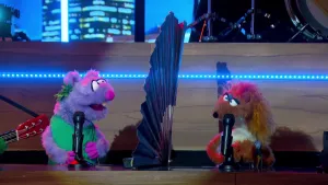 Foto van scène uit de Boterhamshow met Lucy en Roos in de studio aan de desk die allebei microfoon hebben, tussen hen in staat een grote waaier.