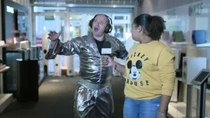 Foto van presentator die man met oortelefoon op en met zilverkleurige kleding aan interviewt.