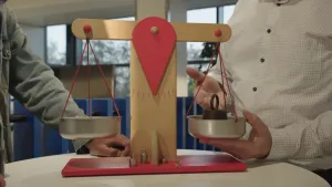 Foto van een houten speelgoedweegschaal met twee schalen, in de ene zit een gewicht van een kilo.