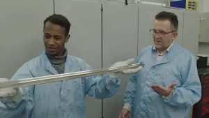 Foto van twee mannen in werkkleding met een metalen staaf van een meter lang in de handen.
