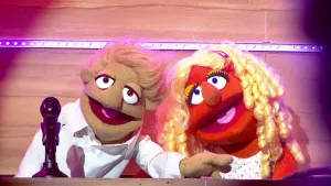 Foto van scène uit de Boterhamshow waarbij man en vrouw (poppen) aan de desk zitten bij een microfoon, zingen een liedje.