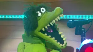 Foto van scène uit de Boterhamshow met de kop van een krokodil (pop) die zingt in microfoon.