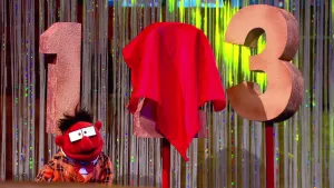 Foto van scène uit de Boterhamshow met Pi R Kwadraat aan tafel, erachter een één, een afgedekt cijfer, een drie.