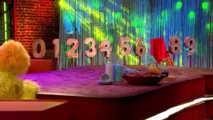 Foto van een scène uit de Boterhamshow waarbij Opperdepop en Pi R Kwadraat aan tafel zitten, reeks cijfers op de achtergrond waarbij de zeven is afgedekt.