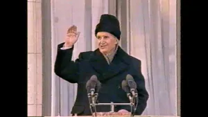 Een foto van de Roemeense dictator Ceausescu die zwaait naar mensen buiten beeld. Hij staat achter een standaard met zes microfoons eraan. Hij draagt een zwarte jas en een zwarte muts op zijn hoofd. Erachter hangen doorzichtige,witte gordijnen, vitrage.