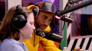 Foto van een meisje achter de microfoon in de studio, erbij zit presentator/DJ.