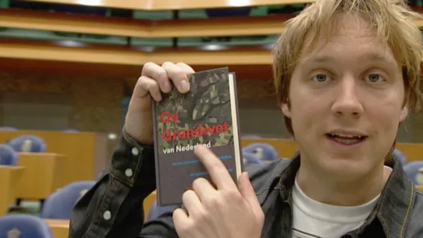 Foto van presentator in de Tweede Kamer die een boek vasthoudt: de Grondwet.