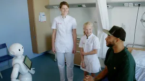 Foto van een ziekenhuiskamer met presentator, een kind en verpleegster en er staat een kleine witte robot.