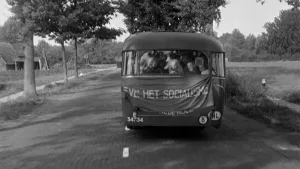 Zwart/wit-foto van een bestelbusje dat op een weg met bomen rijdt met spandoek met tekst aan de voorkant.