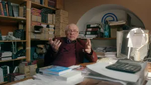 Foto van een man aan tafel met boeken en andere dingen erop, erachter kast met boeken etc. erin.