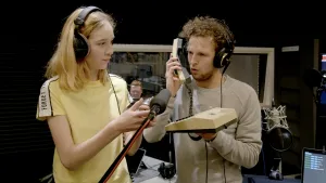 Foto van een meisje en man met beiden oortelefoon op in opnamestudio, de man houdt een telefoon vast bij een microfoon.