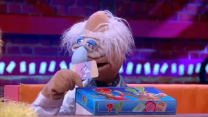 Scène uit de Boterhamshow met Professor Bolleboos (pop) die aan de desk zit met een spelletjesdoos.