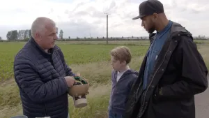 Foto van presentator en jongetje buiten op weiland met een man die een vuurwerkbom (projectiel) in de handen houdt.