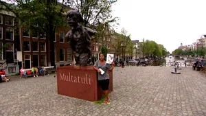 Foto van presentator bij een beeld van Multatuli op straat te Amsterdam, ze houdt een portret van hem vast.