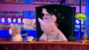 Foto van scène uit de Boterhamshow met Nico de Neushoorn en een mug (poppen) aan tafel, de mug houdt tekening van een arm vast.