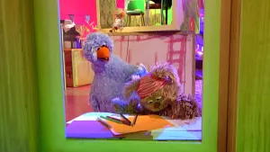 Foto van een scène uit Sesamstraat met Pino en ander Sesamstraatfiguur aan tafel met vellen papier en kleurpotloden.