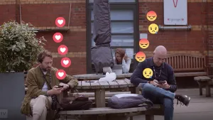 Foto van twee mannen op een bank die appen met elkaar met de telefoon, op het beeld zie je emoticons (hartjes, boze gezichtjes) erbij.