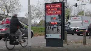 Foto van straatbeeld met auto's en fietser, op de stoep staat groot lichtbord met instructies voor fietsers erop.