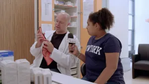 Man, een arts, wordt ondervraagd door presentator in onderzoekskamer, die arts houdt een hand van papier vast.