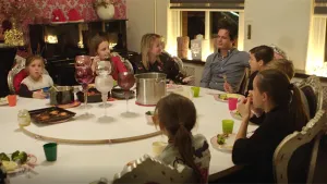 Kinderen zitten samen met een man aan grote tafel in woonkamer met glazen, bekers, een pan etc. erop.