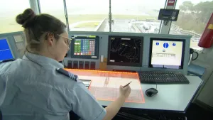Foto van vrouw zit aan tafel met beeldschermen voor raam in verkeerstoren op een vliegveld.