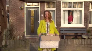 Vrouw staat voor ingang huis met grote doos in haar handen, door het raam van dat huis kijkt een man naar buiten.