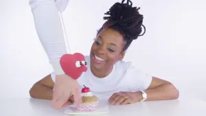 Foto van een meisje dat aan tafel zit en krijgt van iemand taartje aangereikt, aan de mouw van diegene is een stoffen hartje bevestigd.