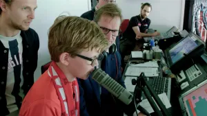 Foto van omroepinstallatie van een stadion waarbij een jongetje iets zegt in de microfoon, verder zitten er volwassen mensen omheen.