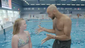 Foto van een meisje in zwemkleding staat samen met een man met ontbloot bovenlijf bij een groot zwembad.