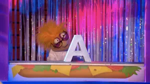 Foto van de Boterhamshow, je ziet Co Sinus (pop) aan tafel waarop een grote letter A staat.