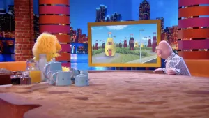 Foto van Boterhamshow, Opperdepop en andere pop (die architect) aan tafel, in het grote scherm aldaar tekening van huizen in de vorm van frisdrankflessen.