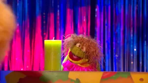 Foto van Boterhamshow waarbij een meisje (pop), Benthe, zit aan tafel waarop ook een grote kaars (niet aan) staat.