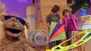 Beeld van uitzending Sesamstraat met Tommie en paar mensen bij sportartikelen zoals badmintonrackets, een vlieger.