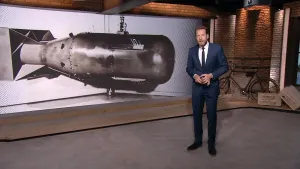 Foto van beeld uit het NOS-Journaal met de nieuwslezer die staat bij een model van een atoombom.