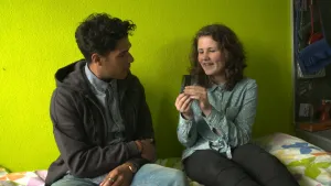 Foto van man en vrouw die zitten op een bed en zijn in gesprek, de vrouw houdt een luizenkam vast.