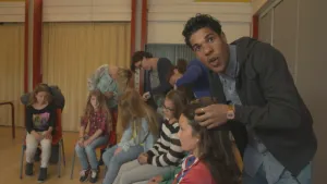 Foto van scène uit Klokhuis met man en vrouw, kinderen zitten erbij, vrouw controleert één kind op hoofdluis, verder de presentator.