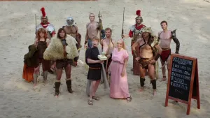 Foto van aantal mensen en kinderen verkleed als Romeinen met wapens zoals een lans, staan op het zand van het Colosseum.