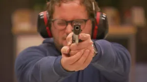 Een foto van Diederik, die een pistool in zijn handen heeft en de loop van het pistool richt naar een punt achter de camera. Op zijn hoofd heeft hij een geluidsbeschermer zitten, zwart met rode randen om de oorstukken heen. 