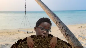 Een foto van een strand met een soort houten schommel aan een kromme boomstam. Vooraan is Jona te zien, waarvan hoofd en handen uit een hoop donkere aarde steken, waardoor het lijkt alsof hij uit de grond gekomen is zoals een mol uit een molshoop. 