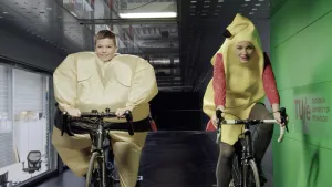 Een foto van Levi en Tess die allebei fietsen in een ruimte van de Technische Universiteit Eindhoven. Levi draagt een sumoworstelaarspak, waardoor het lijkt alsof hij heel dik en breed is. Tess draagt een bananenpak over haar kleding.
