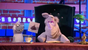 Een foto van pop Stella Schaap, die een wit bord vastheeft met een getekende bruine, gekrulde drol erop. Ernaast zit de pop Nico de Neushoorn op een groene stoel achter een bureau met een opengeslagen boek en een wit theekopje erop. 