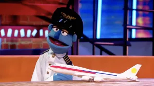 Een foto van blauwe pop Frits-Boudewijn van Laarhoven in pilotenkleding: een witte blouse met goudkleurige knopen en een zwarte pet met goudkleurige vleugels erop. Op de tafel vóór de pop staat een passagiersvliegtuig van wit plastic.