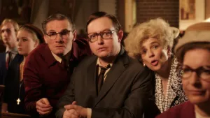 Een foto van mensen die achter een kerkbank staan. Drie mensen, twee mannen in pak en één vrouw in een jurk met een ketting van witte bolletjes om, kijken in de richting van de camera. Drie andere mensen kijken naar een punt links buiten beeld.