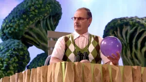 Een foto van Buurman Baasje achter een hek van houten palen. Hij heeft een paarse bal van plastic vast. Hij draagt een zwarte verrekijker om zijn nek. Op de achtergrond staan vier grote, groene dingen in de vorm van de groente broccoli.