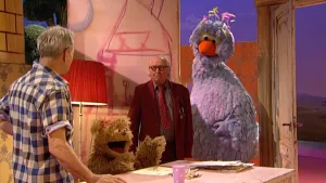 Een foto van Frank die links in beeld van achteren te zien is en kijkt in de richting van Pino en meneer Aart, die rechts in beeld staan. Pino staat naast een geopende deur. Ertussenin staat Tommy achter een tafel vóór een tekening en een stapel krijtjes.