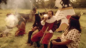 Een foto van een grasveld waar iemand verkleed als Henry Hudson een pijp rookt naast twee Native Americans of Indianen, met een veer om hun hoofd gebonden, waarvan er ook één iets boven een vuur houdt. Een wigwam, een soort puntige tent, staat erachter.