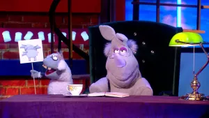 Een foto van de pop Nico de Neushoorn op een groene stoel achter een bureau met een opengeslagen boek. Naast Nico staat de pop Stella Schaap. Stella heeft een bord vast met daarop drie getekende schapen met een witte vacht van wol.