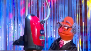 Een foto van pop Hugo Baas die naar een rood met zwarte stofzuiger kijkt die op de verder niet-zichtbare boterhamband staat. De pop heeft een zwart, glimmend pak aan, een witte blouse, een roze stropdas om zijn nek en een bril met bruine panterprint op.