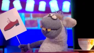 Een foto van pop Stella Schaap. De pop heeft een bord met een getekende rode schoen erop vast en kijkt naar het bord. De pop heeft een witte vacht op haar rug en oren, een grijze vacht bij kop en poten, ronde ogen en vooruitstekende, gele tanden.