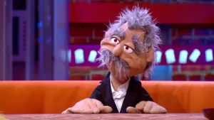 Een foto van pop Kees Kniesoor op een bruine bank achter een bruine tafel. De pop heeft een zwart pak met een witte blouse eronder aan. De pop heeft een lichtbruine huid, grijs haar, dikke grijze wenkbrauwen en een grijze snor. 